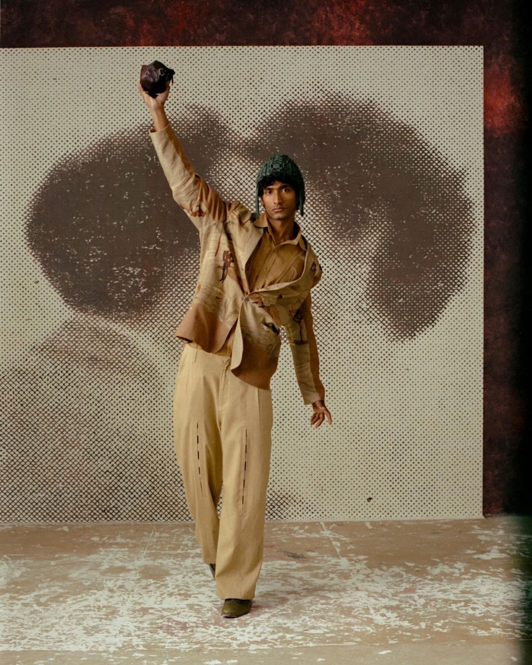 A tan-skinned man in a beige suit raises a rock in a studio setting with a grunge halftone background for a fashion or editorial shot.