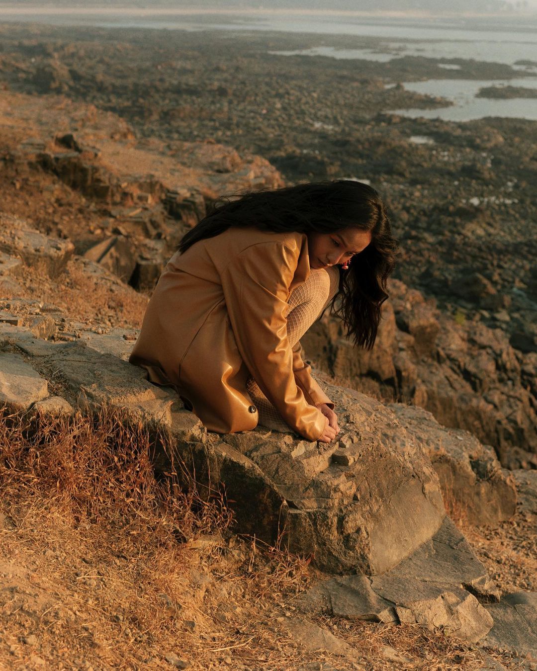 Woman in tan trench coat crouched on rocky coastal terrain overlooking a shoreline by photographer Dhruvin in Mumbai