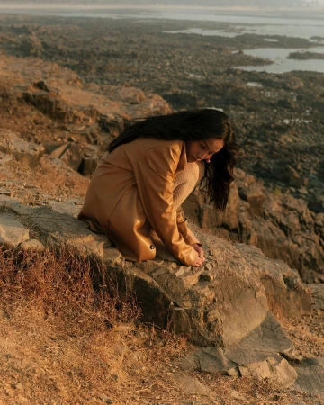 Woman in tan trench coat crouched on rocky coastal terrain overlooking a shoreline by photographer Dhruvin in Mumbai