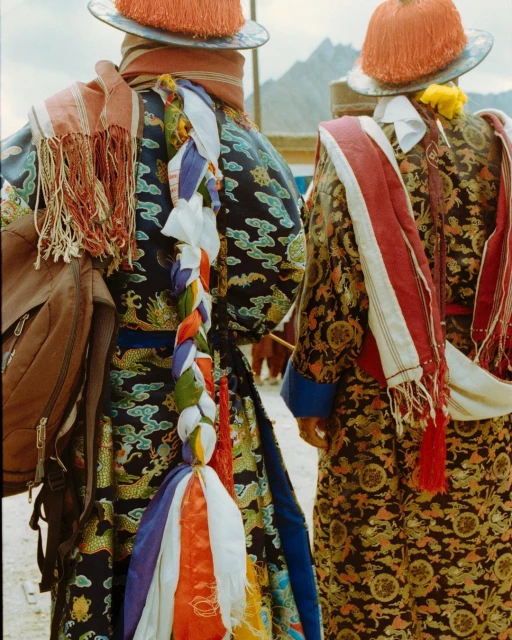 Two people in ornate, traditional clothing and hats stand outdoors against a blurred background of low mountains and sky.