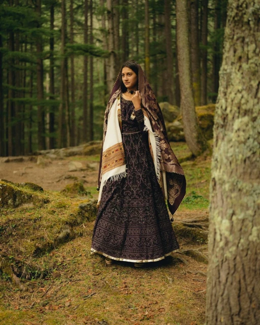 Woman in patterned dress with shawl poses in a forest clearing, surrounded by tall trees and mossy ground in a natural light.