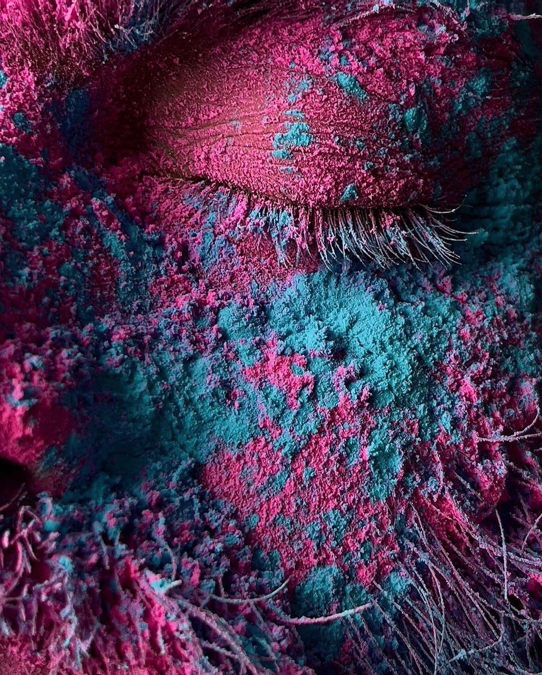Close-up portrait of a face covered in a layer of brightly colored pink and teal powder with visible eyelashes and facial hair.