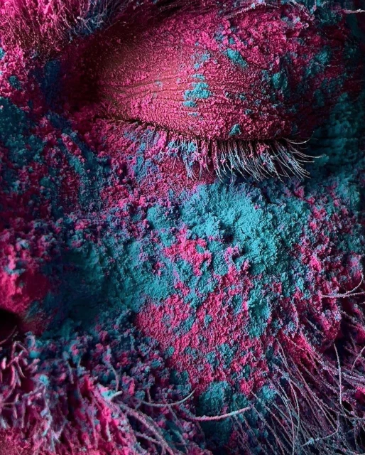 Close-up portrait of a face covered in a layer of brightly colored pink and teal powder with visible eyelashes and facial hair.