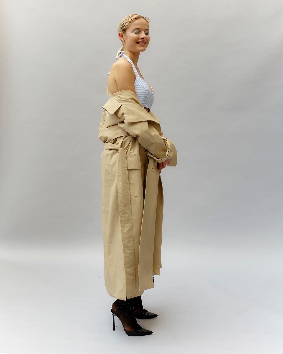 Woman in blue and white halter top and tan trench coat poses in black heels in a photo studio with a light grey backdrop.