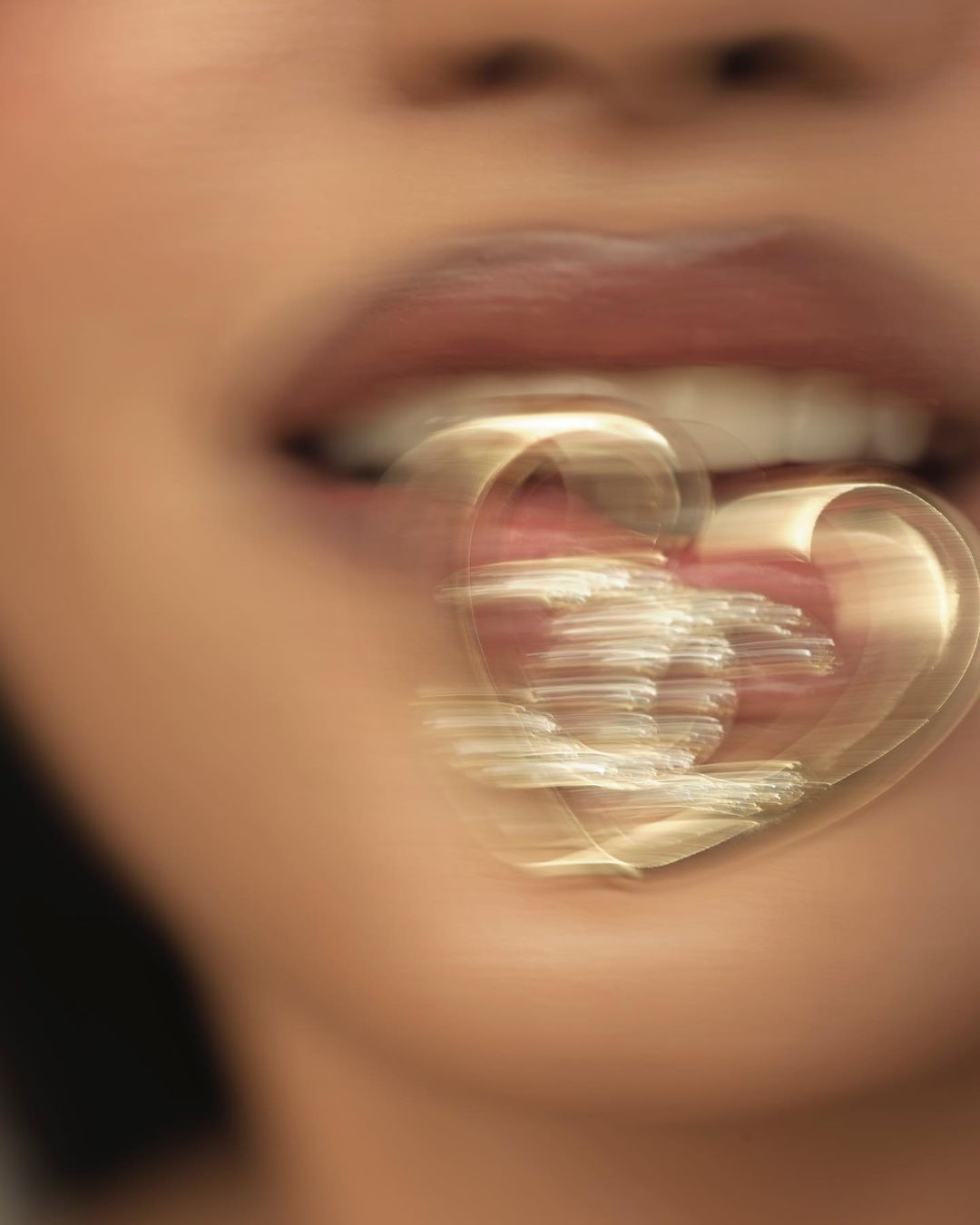Close-up of an adult female's mouth with her lips slightly parted, revealing white teeth, and a blurred golden heart-shaped object.