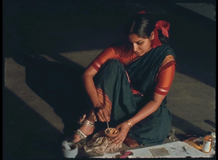 Adult female sitting on floor prepares material in small bowl; mortar and pestle visible, wearing sari and jewelry, with shadows visible.