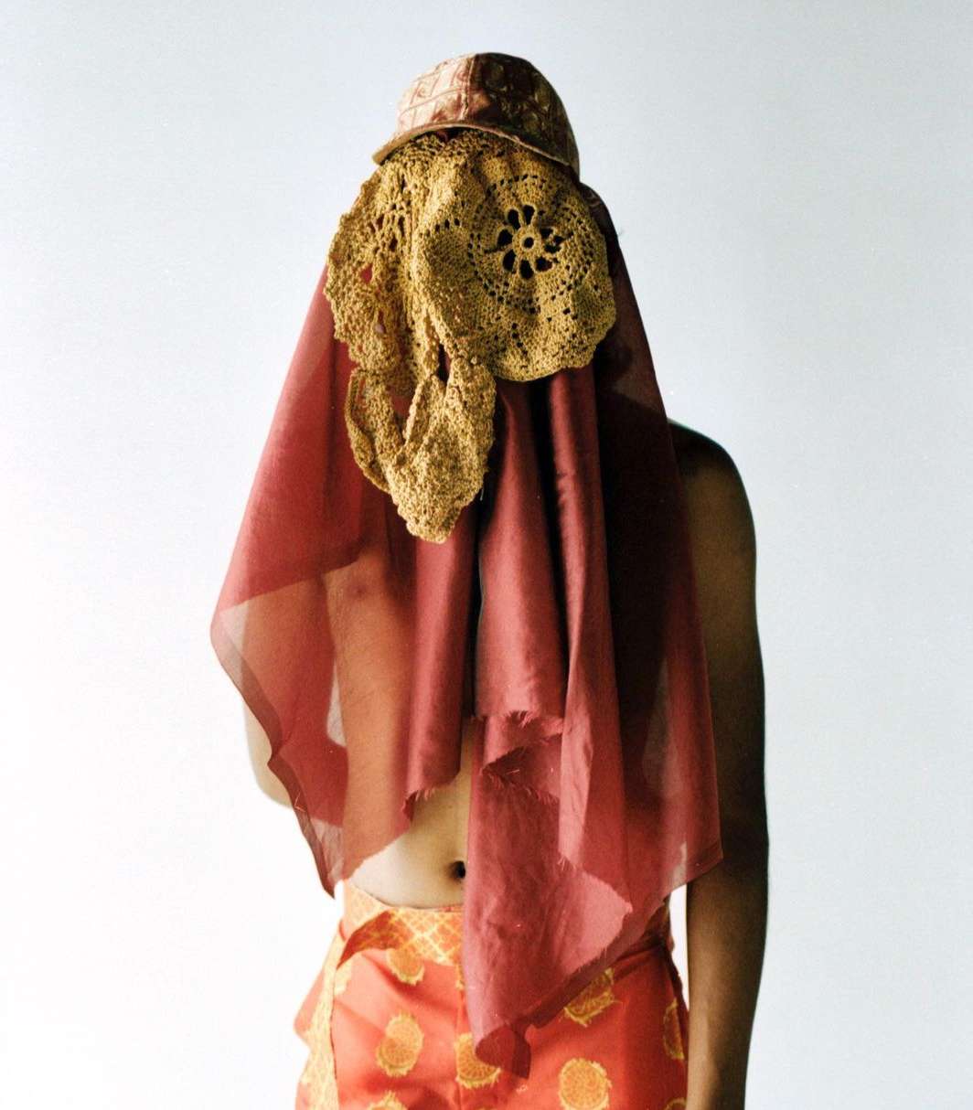 Person with crocheted hat and sheer drape over their upper body, wearing patterned pants, against a white backdrop in a studio.