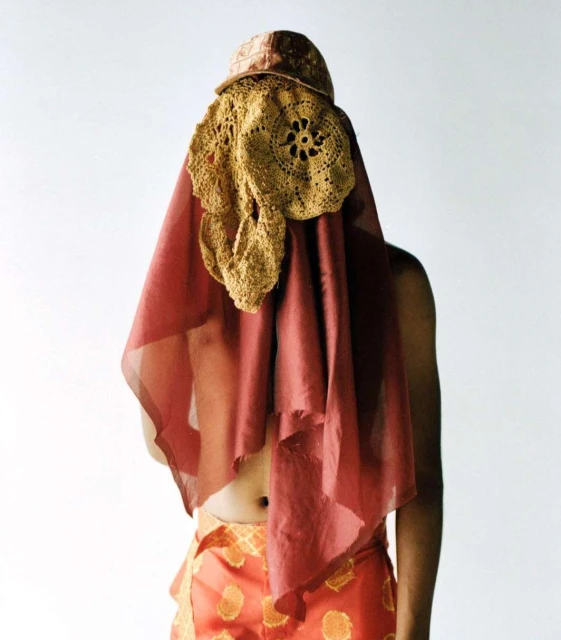 Person with crocheted hat and sheer drape over their upper body, wearing patterned pants, against a white backdrop in a studio.