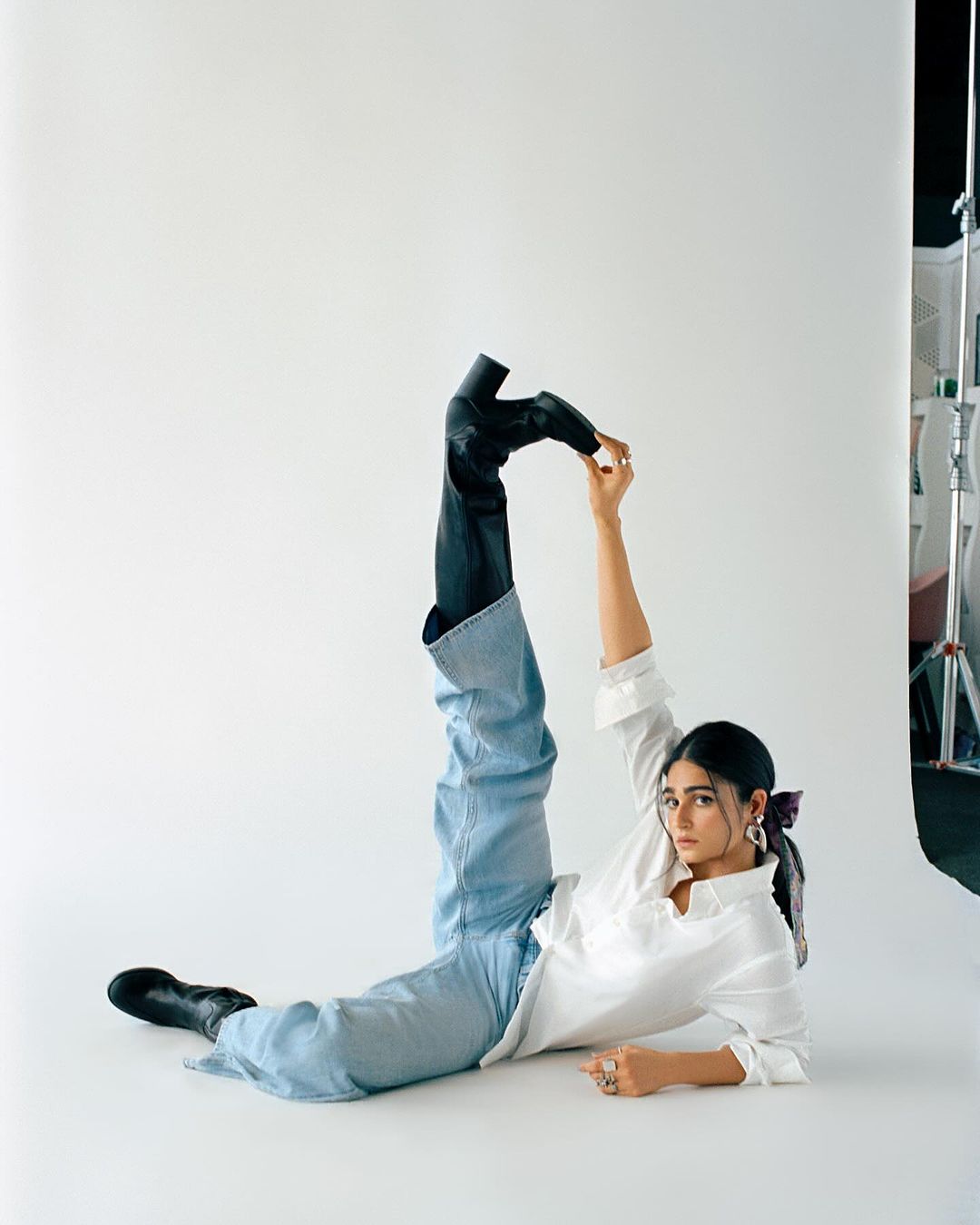 Woman in a white shirt and blue jeans poses in a studio with one leg up in the air, holding her foot wearing a black boot.