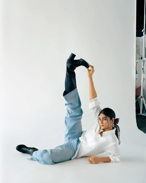 Woman in a white shirt and blue jeans poses in a studio with one leg up in the air, holding her foot wearing a black boot.