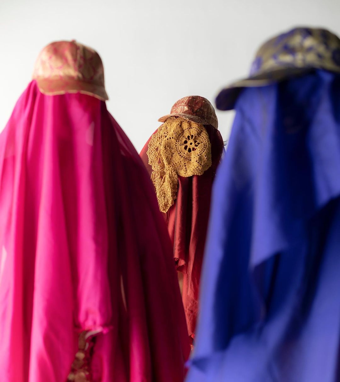 Three veiled figures stand against a white background. The figures are draped in bright pink, red, and blue fabric and topped with patterned caps.