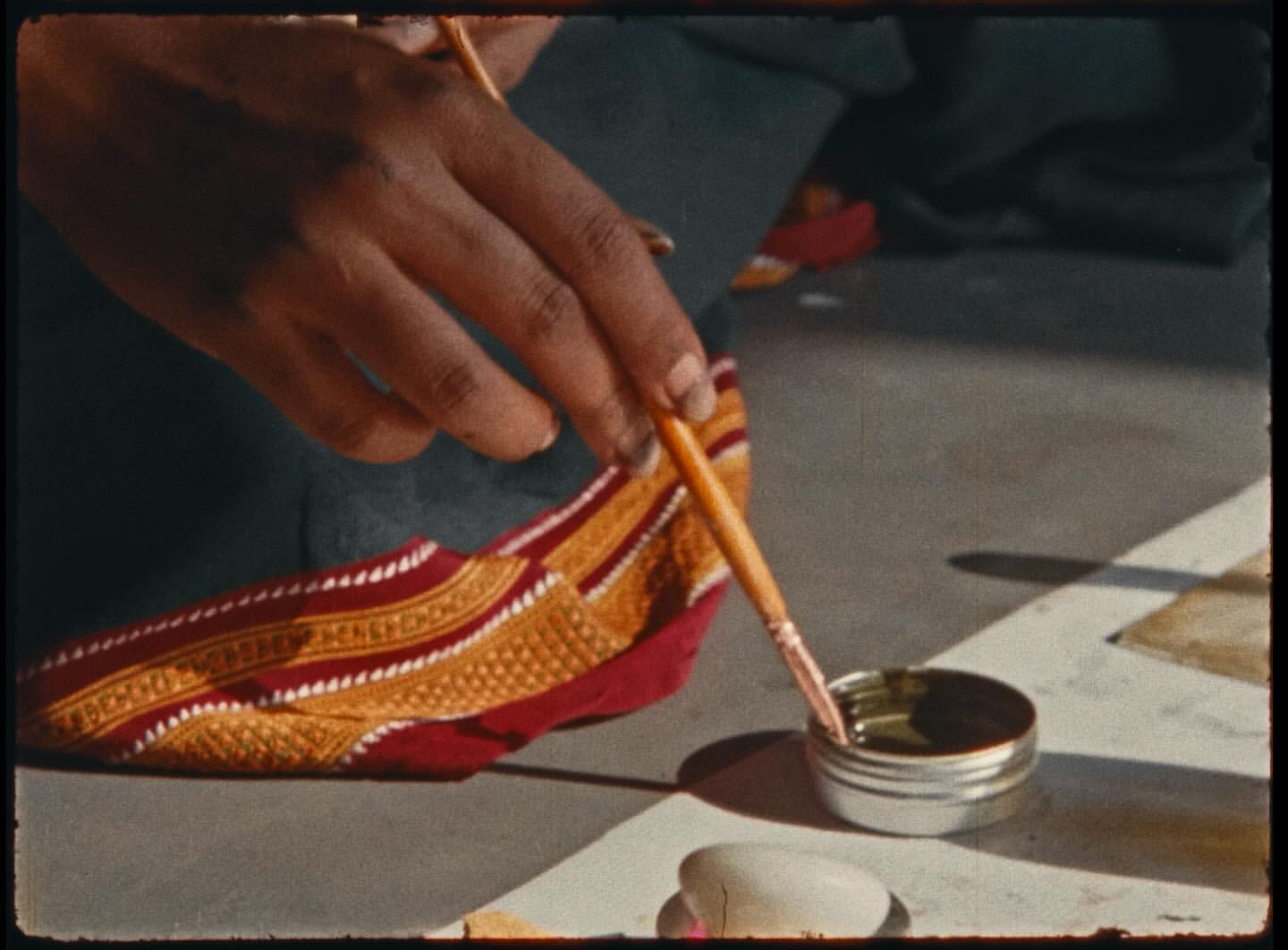 Close-up of a person's hands painting with a brush, focusing on the brush, paint container, and detailed textile design nearby.