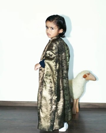 Child wearing a metallic gold long coat looking over shoulder with a toy sheep in the background