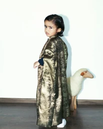 Child wearing a metallic gold long coat looking over shoulder with a toy sheep in the background