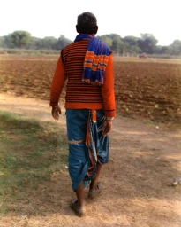 Rear view of an elderly man walking along a dirt path beside a harvested field wearing an orange striped sweater and blue wrap skirt.