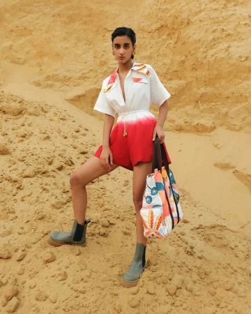 Model standing on a sandy terrain wearing a white and red ombre romper and carrying a patterned tote bag