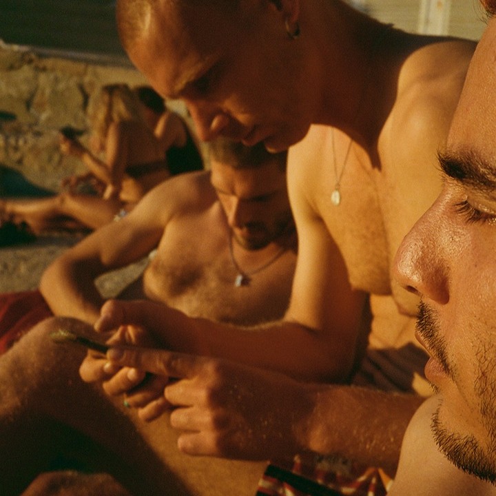 Group of shirtless men using mobile phones on a sunny beach, with warm lighting and a close-up camera angle.