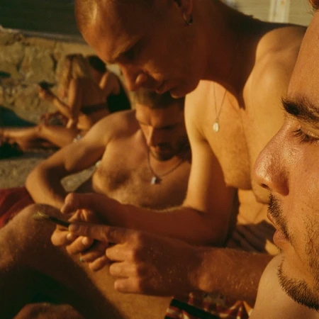 Group of shirtless men using mobile phones on a sunny beach, with warm lighting and a close-up camera angle.