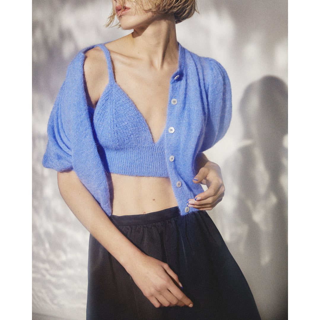 Woman in blue knit bralette, open cardigan, and black satin pants, posing indoors with a soft background and natural lighting.
