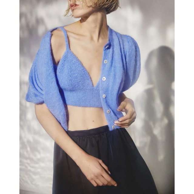 Woman in blue knit bralette, open cardigan, and black satin pants, posing indoors with a soft background and natural lighting.