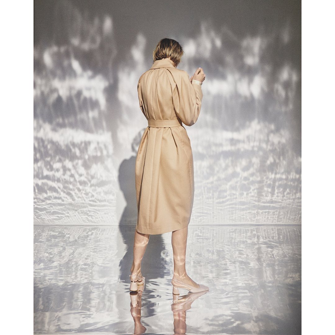 Back view of an adult woman wearing a beige trench coat and matching boots posing in a studio setting with dappled light.