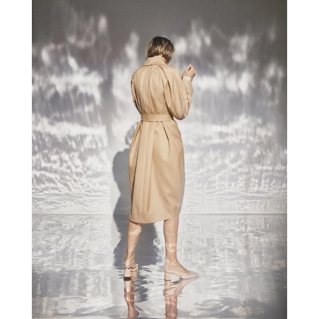 Back view of an adult woman wearing a beige trench coat and matching boots posing in a studio setting with dappled light.