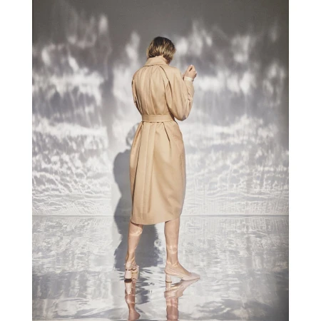Back view of an adult woman wearing a beige trench coat and matching boots posing in a studio setting with dappled light.