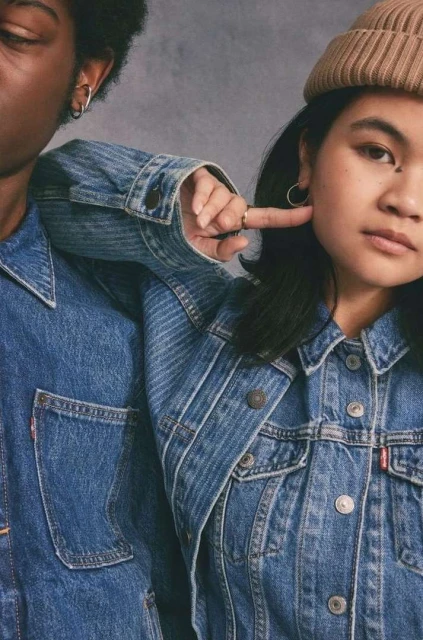 Two people wearing denim jackets in front of a grey background. The person on the right is pointing at their face.