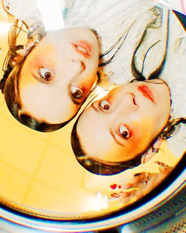 Upside-down portrait of two young adult women with elaborate makeup and jewelry, reflected in a gold surface with studio lighting.