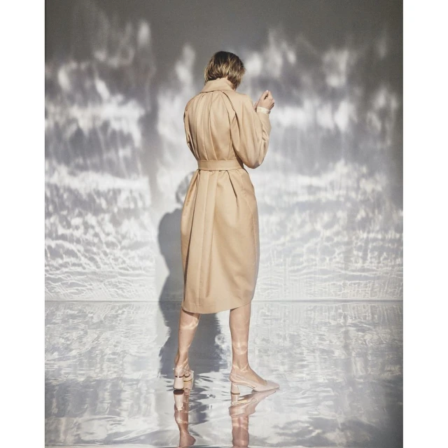 Back view of a woman in a tan dress with matching boots, standing indoors with dappled light patterns on the wall behind her.