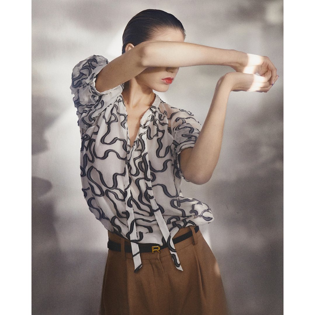Fashion shot of a woman with slicked-back hair wearing a patterned blouse and brown slacks, posing in front of a grey background.