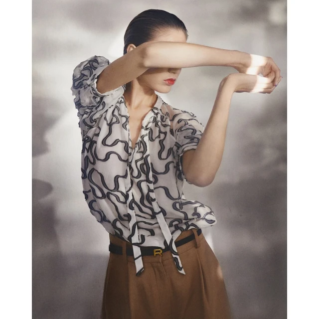 Fashion shot of a woman with slicked-back hair wearing a patterned blouse and brown slacks, posing in front of a grey background.