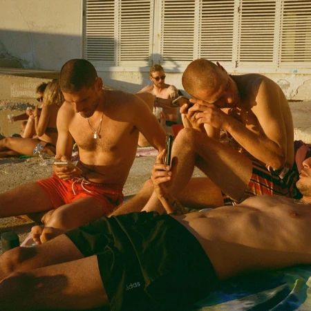 Several adults gather on a sunlit beach, some seated, others lying down, all seemingly engrossed in their smartphones in an outdoor setting.
