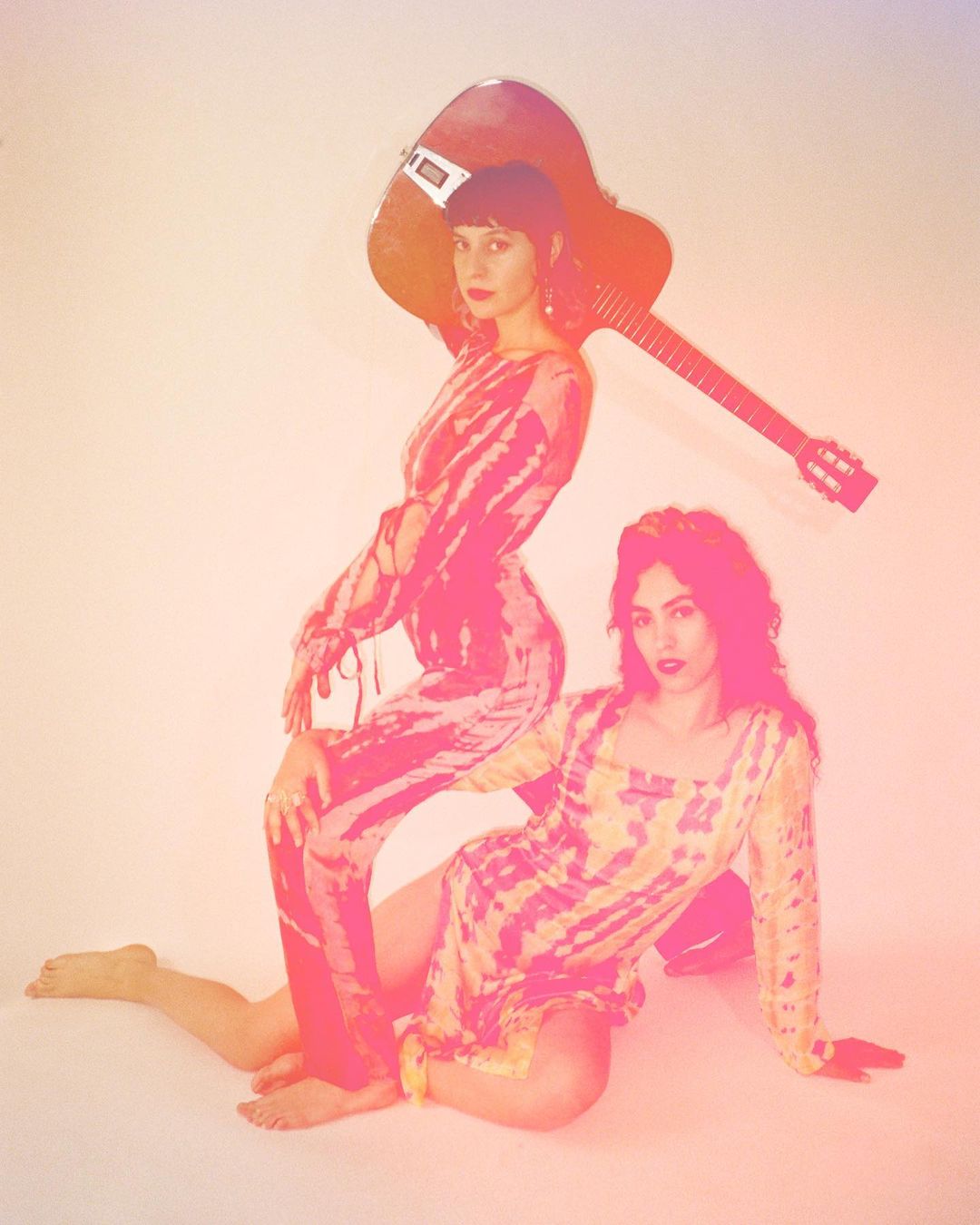 Two women pose in pink-toned studio lighting. One woman stands holding a guitar, and the other sits on the floor in a short dress.