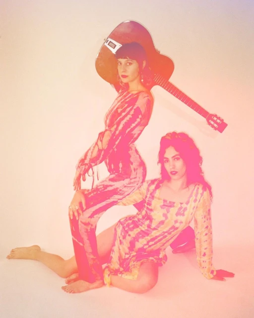 Two women pose in pink-toned studio lighting. One woman stands holding a guitar, and the other sits on the floor in a short dress.