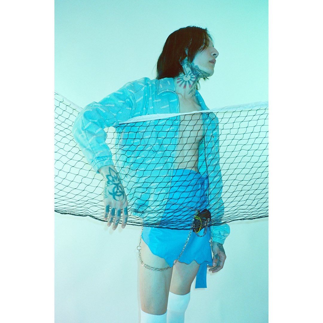 Person with face and neck tattoos wears a blue outfit and white socks, standing in front of a tennis net in a brightly lit studio.