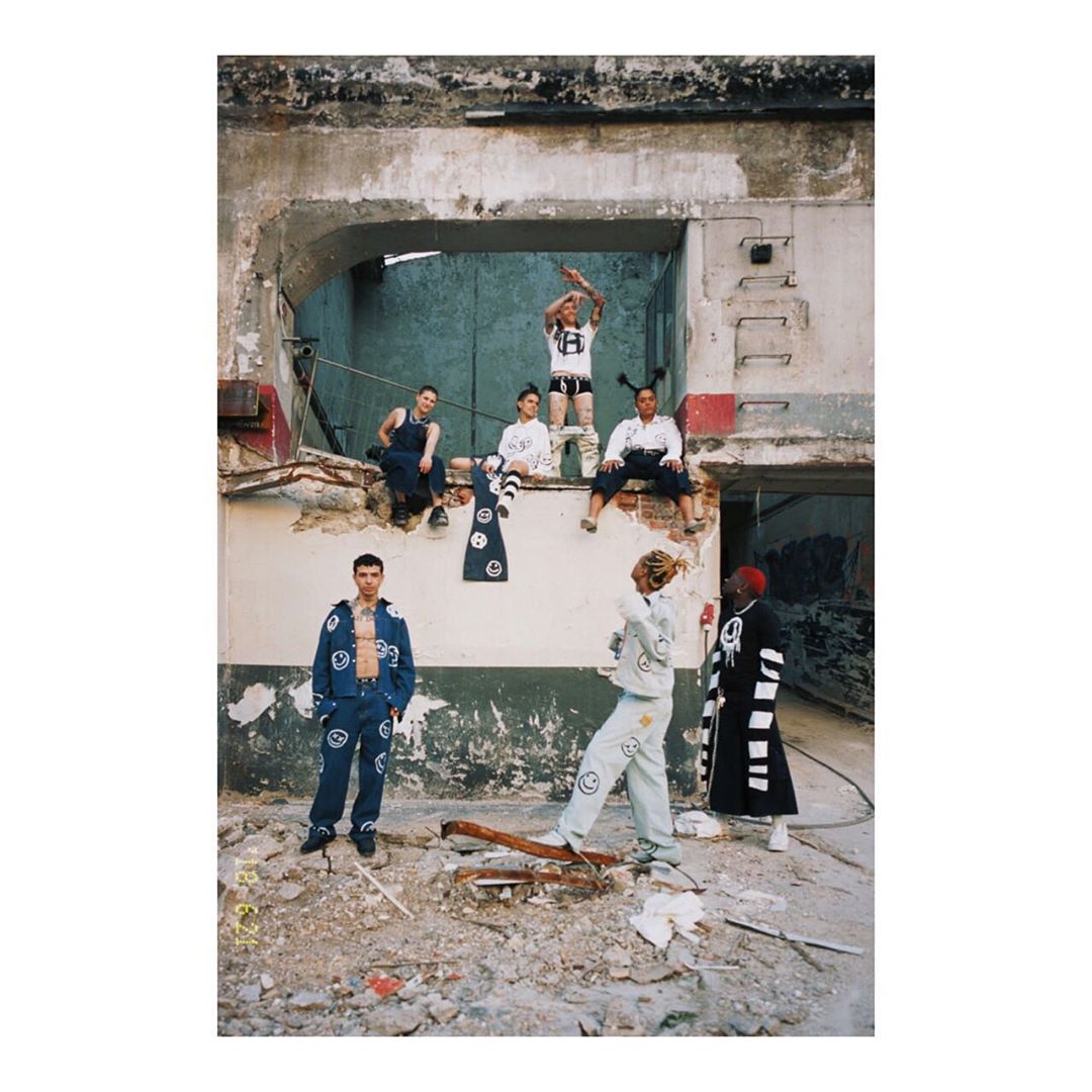 Seven people pose in an urban decay environment; fashion photoshoot; trendy clothing; graffiti and brick background; eye-level shot.