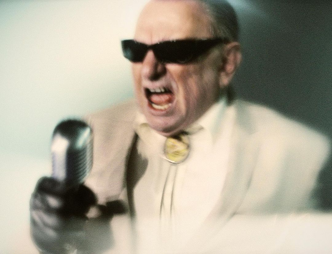 An adult male vocalist wearing dark glasses sings into a microphone, illuminated by a bright light source. The man wears a bolo tie.