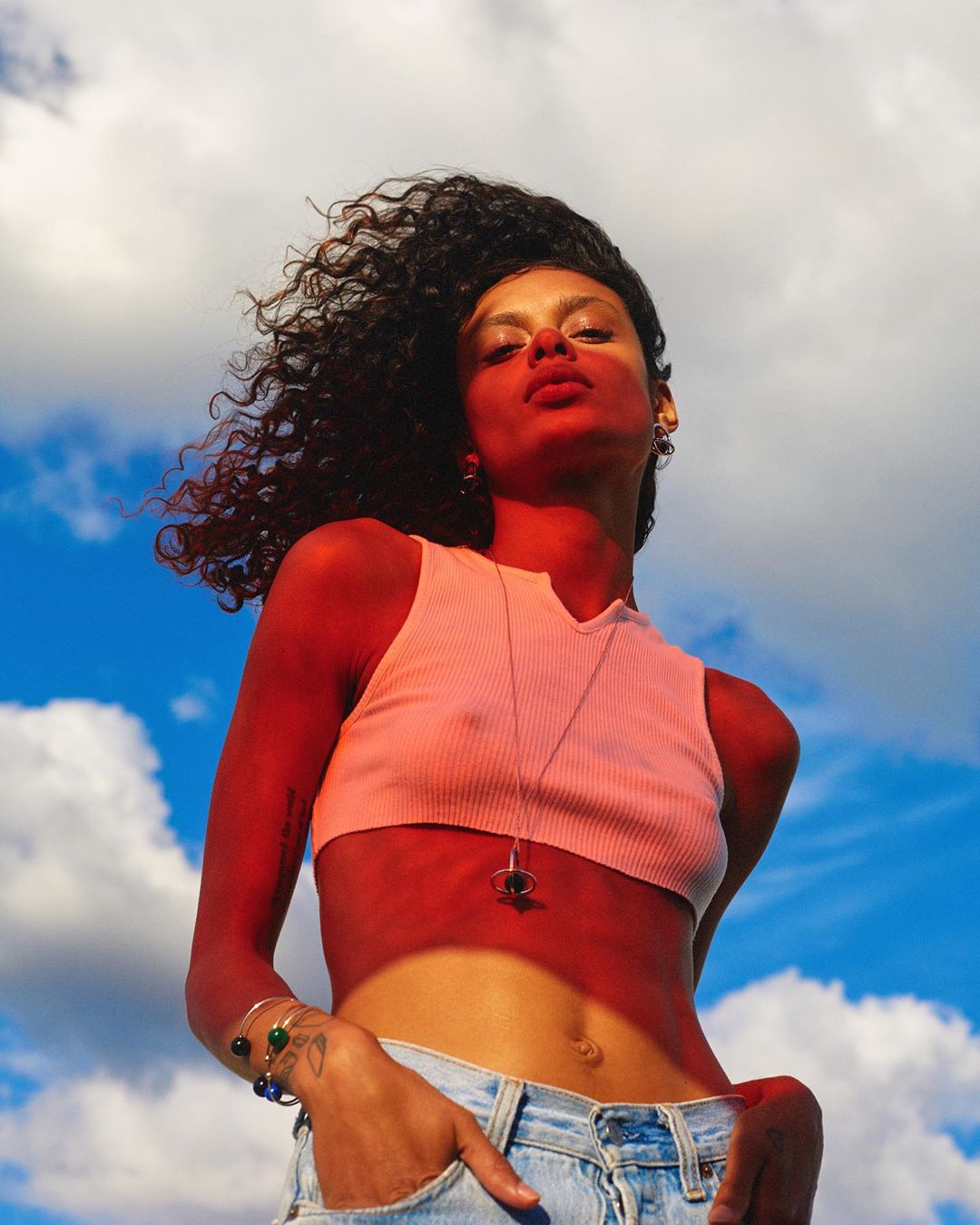 Portrait of an adult female with curly hair, wearing a crop top and jeans, posing in front of a blue sky with white clouds.