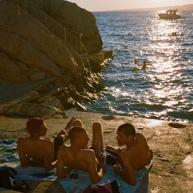 Four young adults sit on a concrete platform near the sea, illuminated by sunlight, with a small boat visible in the distance.