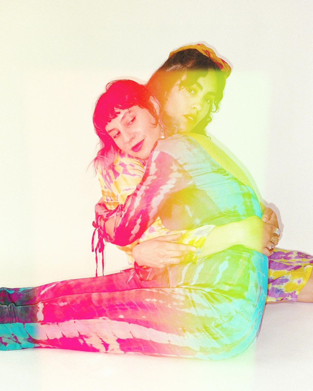 Two women embracing indoors with tie-dye clothing, sitting, and against a neutral backdrop in a colorful portrait. Friendship.