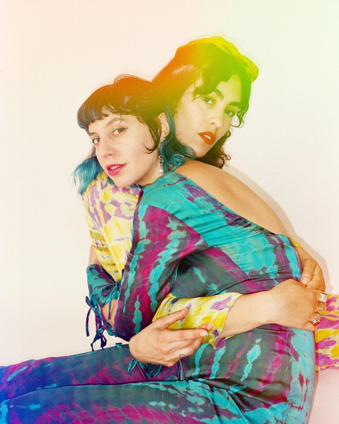 Two adult women pose indoors in colorful tie-dye outfits. One woman embraces the other from behind. Both are looking at the camera.