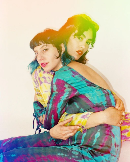 Two adult women pose indoors in colorful tie-dye outfits. One woman embraces the other from behind. Both are looking at the camera.