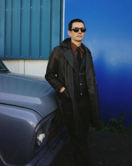 Adult male wearing sunglasses, a dark leather coat, and suit, leaning against a light blue truck with a solid blue wall behind him.