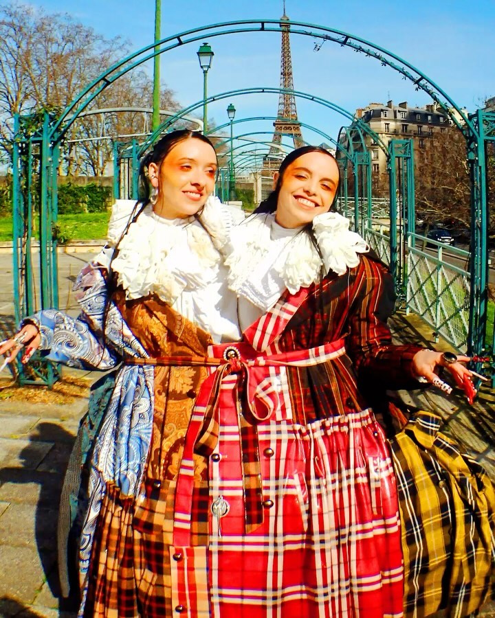 Two young women in ornate costumes pose in front of the Eiffel Tower in Paris. They share one garment with a red plaid skirt.