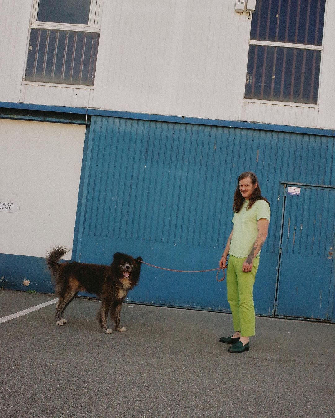 Adult stands with brown, medium-sized mixed-breed dog on leash outdoors in dappled sunlight near a building with barred windows.