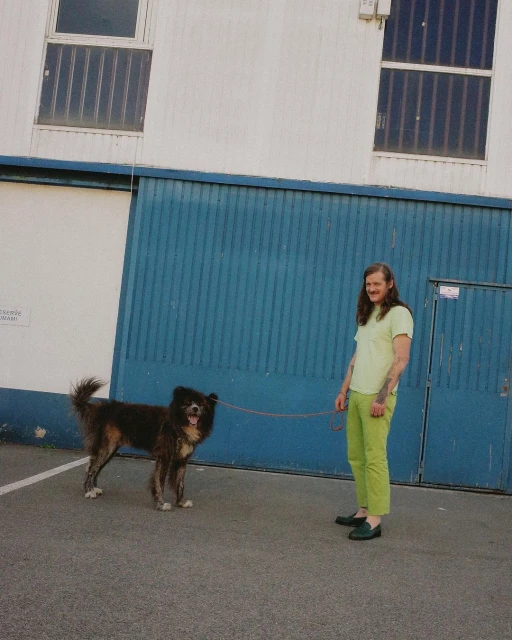 Adult stands with brown, medium-sized mixed-breed dog on leash outdoors in dappled sunlight near a building with barred windows.