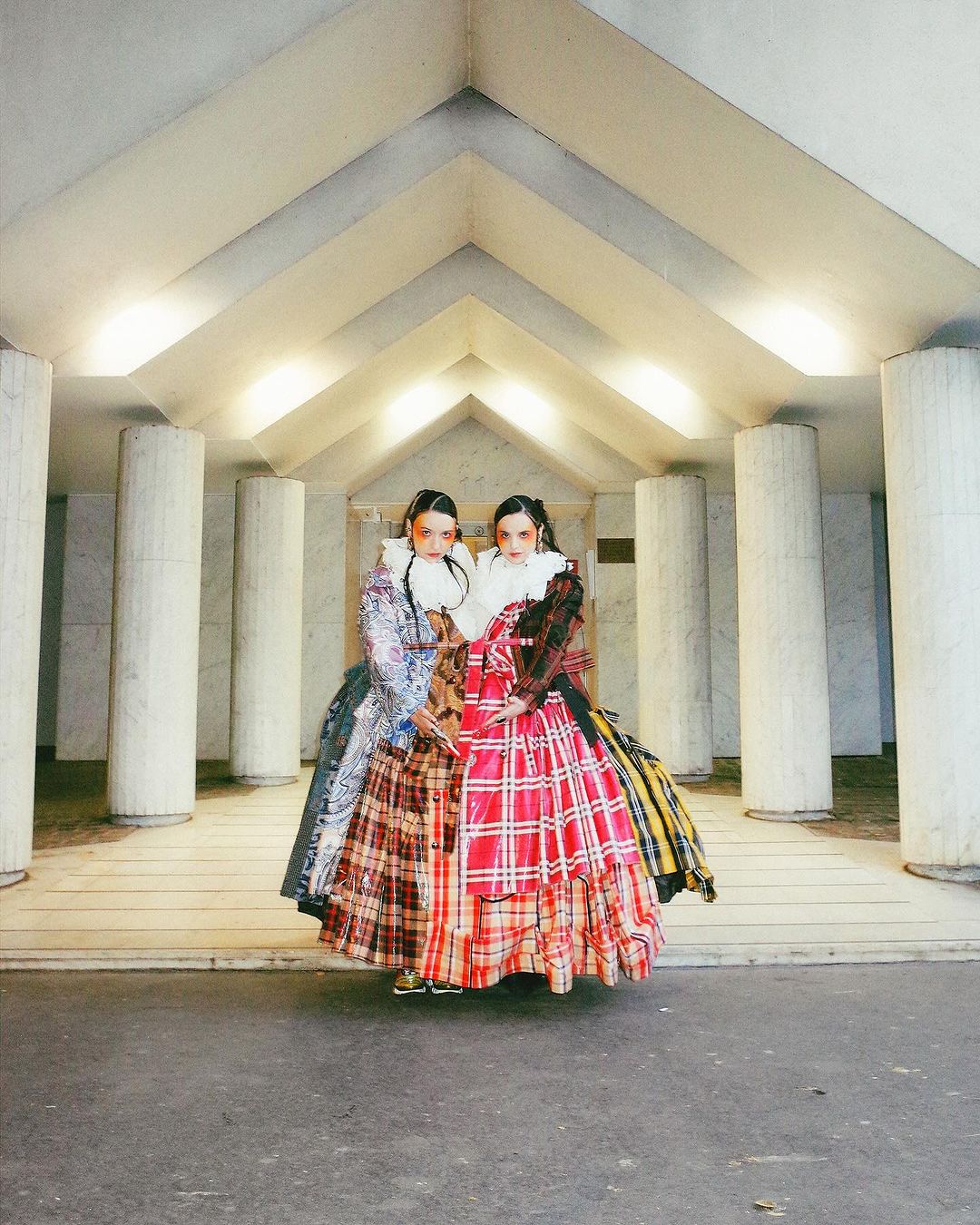 Two women in ruffled collars and plaid gowns stand in an indoor space framed by columns and a modern archway, fashion editorial.