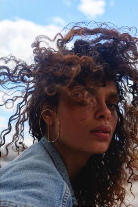 Portrait of a woman with curly hair wearing a gold hoop earring and a denim jacket, posing in front of a blue sky with light clouds.