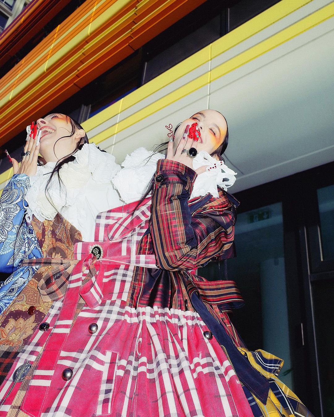 Two women in colorful, patterned dresses stand close together with smiles and exaggerated makeup against an abstract background.